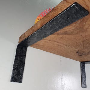 Könnte beinhalten: Ein Holzregal mit natürlicher Holzmaserung, das von zwei schwarzen Metallhalterungen getragen wird. Die Halterungen sind L-förmig und an der Unterseite des Regals befestigt. Das Regal ist an einer weißen Wand montiert.