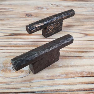 May include: Two rustic metal cabinet door pulls with a hammered finish. The pull on the left is a dark bronze color, while the pull on the right is a black iron color.