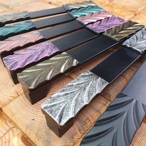 Puede incluir: Una colección de tiradores de cajón con una barra rectangular negra mate unida a una pieza final texturizada con patrón de hoja. Los detalles de las hojas tienen acabados en varios colores metálicos, incluyendo tonos plata, oro, rosa, verde y cobre.