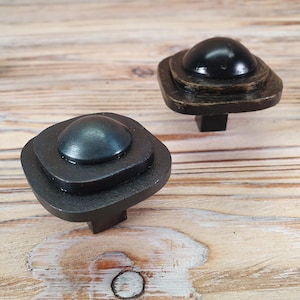 May include: Two black square cabinet knobs with a rounded top. The knobs have a layered design with a raised centre.