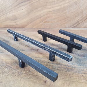 Könnte beinhalten: Drei schwarze Metall-Schrankgriffe mit einer abgenutzten Oberfläche. Die Griffe sind unterschiedlich lang und haben eine rechteckige Form.