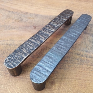 Może przedstawiać: Dwa teksturowane metalowe uchwyty do szafek. Jeden jest w kolorze brązu, drugi w kolorze ciemnoszarym. Oba mają prostokątny drążek z zaokrąglonymi końcami i cylindrycznymi słupkami. Uchwyty są prezentowane na drewnianej powierzchni.