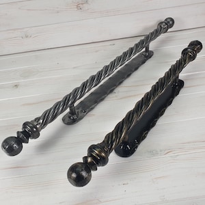 Könnte beinhalten: Zwei schwarze Metall-Türgriffe mit einem gedrehten Seil-Design. Die Griffe haben an jedem Ende einen runden Knopf und sind auf einer flachen Metallplatte montiert.