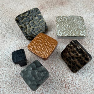 Könnte beinhalten: Eine Sammlung von sechs quadratischen Schrankknöpfen mit unterschiedlichen Texturen und Oberflächen. Das Set enthält Stücke mit dunklem Leopardenmuster, metallischem Silber, strukturiertem Kupfer, tiefem Schwarz und dunkelgrauen Hammerschlag-Oberflächen.