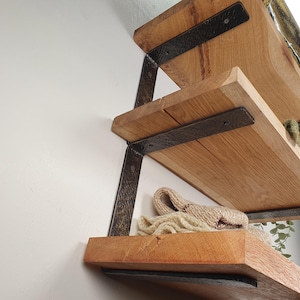 May include: Three-tiered wooden shelves with dark metal brackets. The shelves are made of light-coloured wood and have a natural grain. A beige knitted item rests on the bottom shelf. The brackets are textured and add an industrial touch.