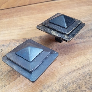 May include: Two square-shaped cabinet knobs with a pyramid-shaped top. The knobs are made of metal and have a dark gray finish. The top knob has a slightly worn finish.