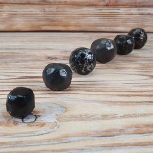 May include: A row of six black, faceted, round metal balls on a wooden surface. The balls are arranged in a semi-circle, with the largest ball at the bottom of the image.