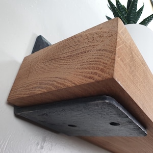May include: A wooden shelf with a natural wood grain finish, supported by a dark gray metal bracket. The shelf is mounted on a white wall. A white pot with a green plant is on the shelf.