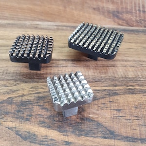 May include: Three square cabinet knobs with a textured, geometric design. The knobs are in black, silver, and gold finishes.