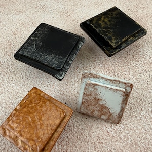 May include: Four square cabinet knobs with a tiered, stepped design. One knob is black with silver distressing, one is black with gold distressing, one is a warm copper tone with a textured finish, and one is white with a copper-toned distressed finish.