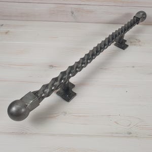 May include: A twisted metal handrail with rounded ends. The handrail is made of dark gray metal and has a rustic, industrial look.