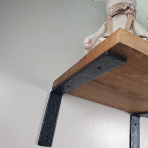 Peut inclure: Une étagère en bois avec une finition naturelle est soutenue par deux supports métalliques noirs texturés. Les supports sont montés sur un mur blanc. Une peluche repose sur l'étagère.