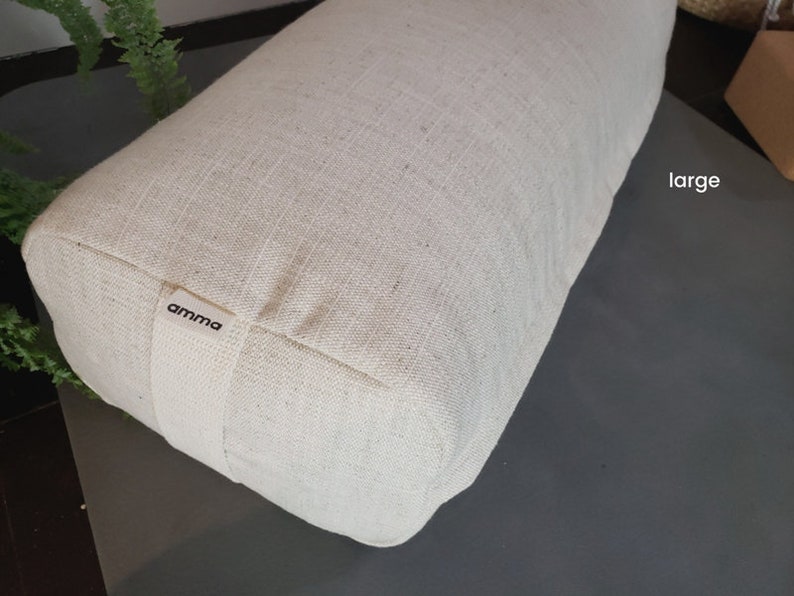 May include: A large, off-white, rectangular meditation cushion with a "amma" label. The cushion is made of a natural fabric and is sitting on a gray surface.