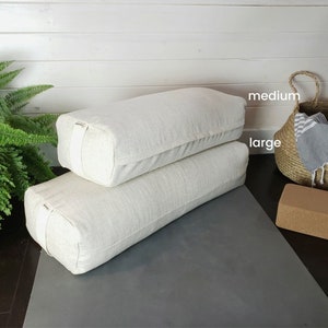 May include: Two white linen yoga bolsters, one labeled "large" and the other labeled "medium". The bolsters are on a gray yoga mat with a cork yoga block.
