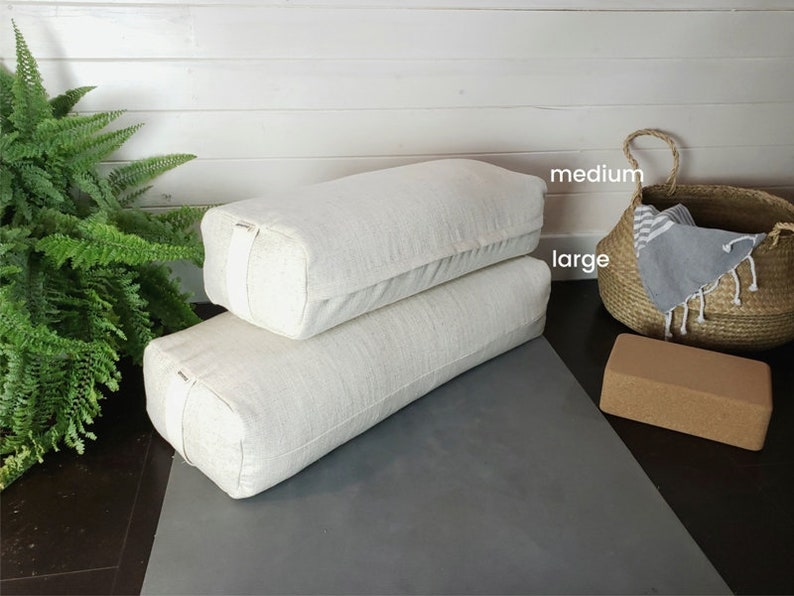 May include: Two white linen yoga bolsters, one labeled "large" and the other labeled "medium". The bolsters are on a gray yoga mat with a cork yoga block.