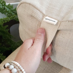May include: A beige fabric cushion with a cream-colored strap and the word "amma" printed on a label. The cushion has a textured surface.