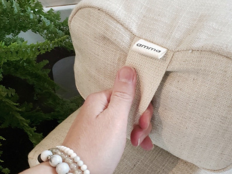 May include: A beige fabric cushion with a cream-colored strap and the word "amma" printed on a label. The cushion has a textured surface.