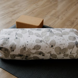 May include: A white yoga bolster with a grey leaf pattern on a black yoga mat. A cork yoga block is in the background.