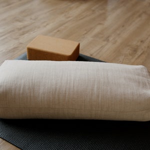 May include: A light beige yoga bolster with a label on the side, resting on a black yoga mat. A brown cork yoga block is in the background.
