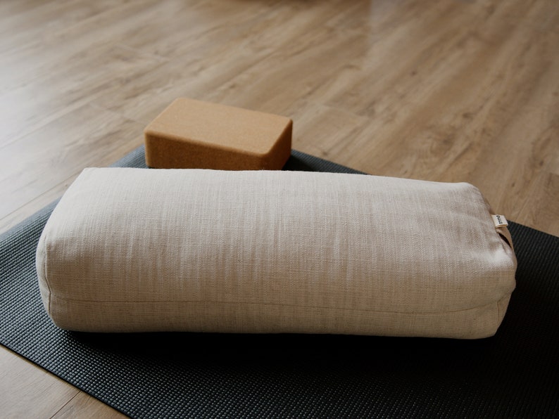 May include: A light beige yoga bolster with a label on the side, resting on a black yoga mat. A brown cork yoga block is in the background.