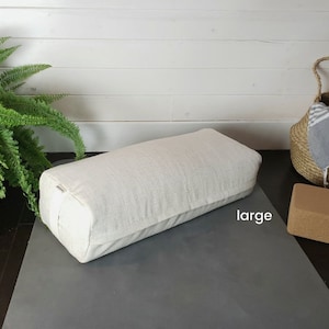 May include: A large, white, rectangular yoga bolster on a gray yoga mat. The bolster is made of a soft, textured fabric and has a white strap. The word "large" is written on the mat.