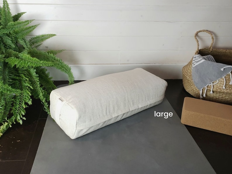 May include: A large, white, rectangular yoga bolster on a gray yoga mat. The bolster is made of a soft, textured fabric and has a white strap. The word "large" is written on the mat.