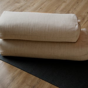 May include: Two beige yoga bolsters stacked on top of each other on a black yoga mat. The bolsters are made of a woven fabric.