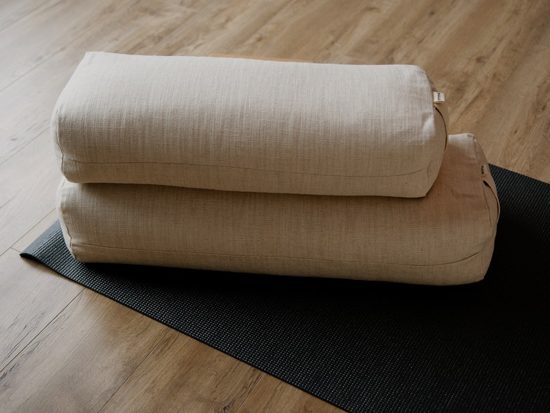 May include: Two beige yoga bolsters stacked on top of each other on a black yoga mat. The bolsters are made of a woven fabric.