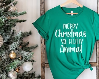 Könnte beinhalten: Ein grünes T-Shirt mit weißem Text, der "Frohe Weihnachten Ya Filthy Animal" lautet. Das Shirt hängt an einer Holzleiter vor einem Weihnachtsbaum.