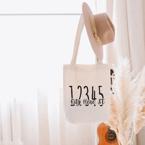 Op de afbeelding: Witte canvas draagtas met zwarte tekst die "12345 City, Here, St" luidt. De tas hangt aan een wit touw en een bruine hoed hangt aan het touw boven de tas. Op de achtergrond staat een boeket gedroogd pampasgras.