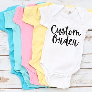 May include: Five baby onesies in different colors, stacked on top of each other. The onesie on top is white with black text that says "Custom Order".