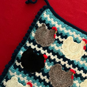May include: Crocheted potholder featuring a blue, white, and navy chevron pattern. The potholder is adorned with several crocheted chicken appliqués in various colors, including white, black, and gray, each with a red comb. A navy blue loop is attached for hanging.