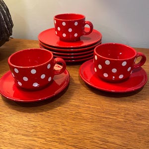 May include: A set of three bright red ceramic cups and matching saucers, each featuring a repeating pattern of white polka dots. The cups have a rounded shape with a side handle, and the saucers are circular with a smooth, glossy finish.