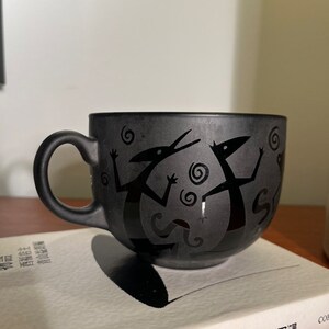 May include: Black ceramic mug with a stylized design of two figures dancing. The figures are black and the background is a dark gray.