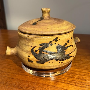 May include: A ceramic lidded jar with a tan base and lid, featuring black abstract designs. The jar has two small handles and a small knob on the lid. It sits on a clear, round stand.