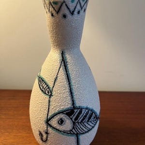 May include: White ceramic vase with a textured surface and a black and blue fish design. The vase has a narrow neck and a wide base.