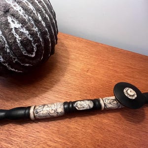 May include: A long, cylindrical smoking pipe featuring alternating segments of smooth black wood and cream-colored material with intricate black line drawings of figures. The pipe includes a circular black bowl with a small, central cream-colored inlay.
