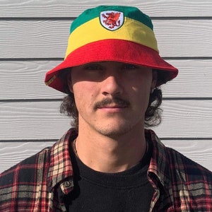 May include: A red, yellow, and green bucket hat with a dragon emblem. The hat is worn by a person with a mustache.