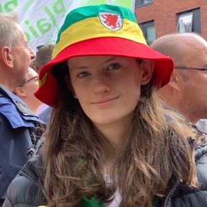 May include: A young person wearing a red, yellow, and green bucket hat with a red dragon emblem. The hat has a yellow band and a green brim.