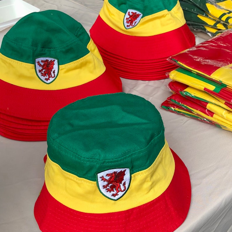 May include: A stack of green, yellow, and red bucket hats with a dragon emblem. The hats are made of a soft, cotton fabric.