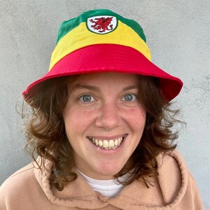 May include: A red, yellow, and green bucket hat with a red dragon embroidered on the front. The hat is worn by a person with curly brown hair.