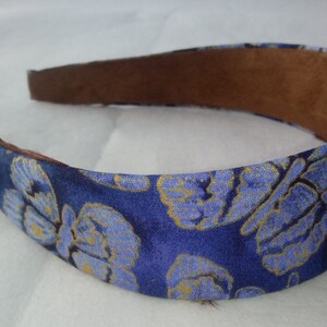 May include: A blue headband with a floral pattern in light blue and gold. The interior is lined with brown material. This hair accessory is suitable for various occasions.
