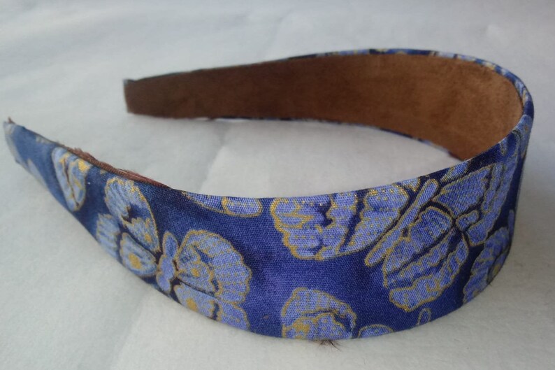 May include: A blue headband with a floral pattern in light blue and gold. The interior is lined with brown material. This hair accessory is suitable for various occasions.