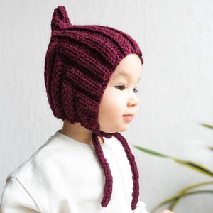 May include: A burgundy knitted baby bonnet with a pointed top and long ties. The bonnet is worn by a baby with a white shirt.