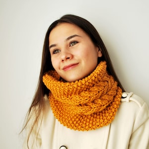 May include: A woman wearing a white coat with a mustard yellow knitted infinity scarf. The scarf has a cable knit pattern.