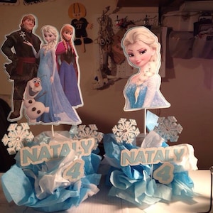 May include: Two Frozen themed centerpieces with blue and white tissue paper, snowflakes, and cardboard cutouts of characters from the movie. The centerpieces are labeled "NATALY 4".