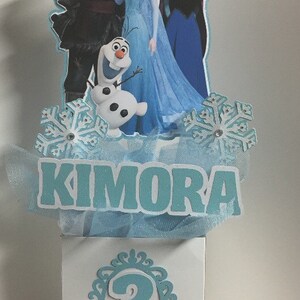 May include: A Frozen themed centerpiece for a birthday party. The centerpiece features a blue, white, and silver snowflake design with a blue tulle bow. The centerpiece also features a cutout of Elsa, Anna, Kristoff, and Olaf from the movie Frozen. The centerpiece has a white box with the number 3 and the name Kimora on it.