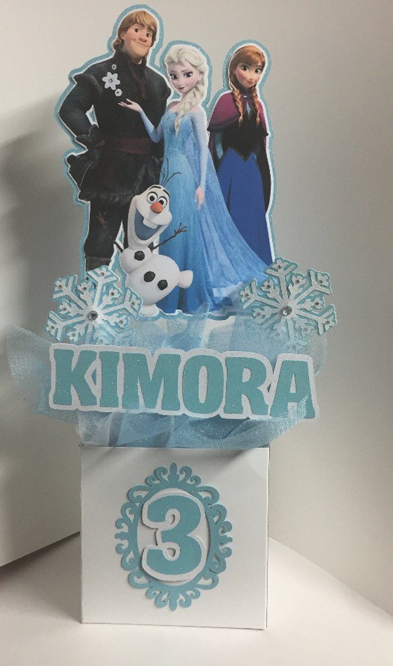May include: A Frozen themed centerpiece for a birthday party. The centerpiece features a blue, white, and silver snowflake design with a blue tulle bow. The centerpiece also features a cutout of Elsa, Anna, Kristoff, and Olaf from the movie Frozen. The centerpiece has a white box with the number 3 and the name Kimora on it.
