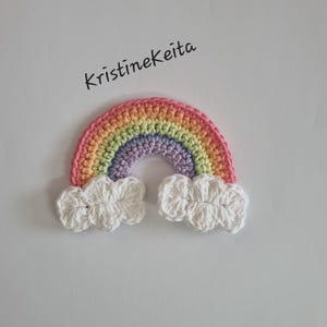 Può includere: Un arcobaleno all'uncinetto con un arco rosa, arancione, giallo, verde e viola e due accenti di nuvole bianche. Il testo "Kristinekeita" è scritto in corsivo nero sopra l'arcobaleno.