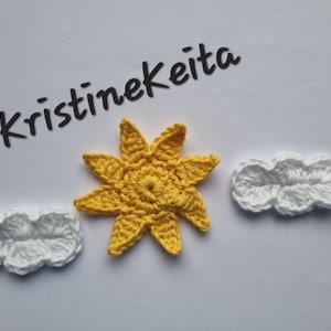 May include: Crocheted yellow sun and two white clouds on a white background. The sun has eight points and a circular center. The clouds are puffy and three-dimensional. The text "Kristinekeita" is above the sun.